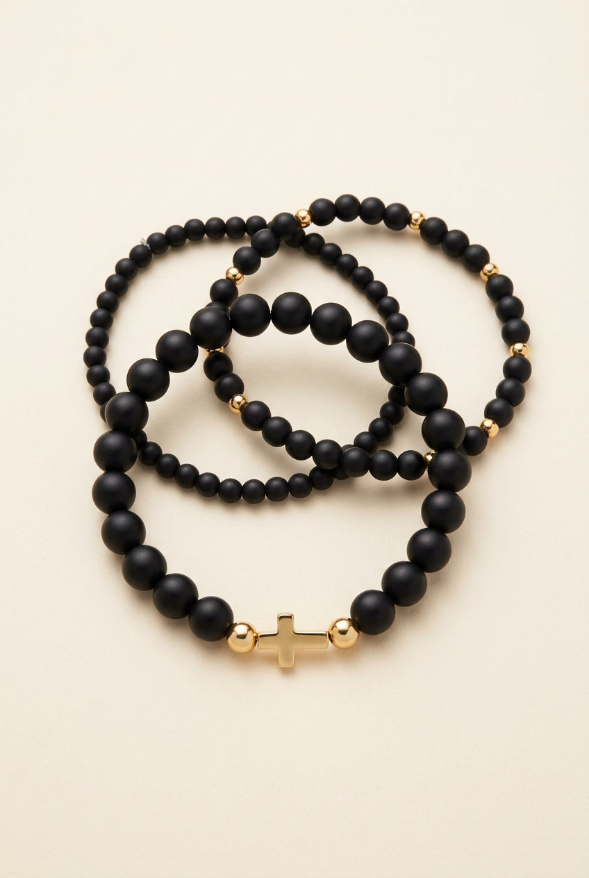 Three black onyx bracelets with gold-colored accents and a cross on a warm ivory background