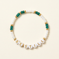 Be Still handmade bracelet with milky white and deep teal-green beads, gold-tone spacers, and gold lettering on an ivory background