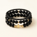 Three handmade black Onyx bracelets arranged in a three-tier stack with gold colored accents and a gold colored cross on an ivory background