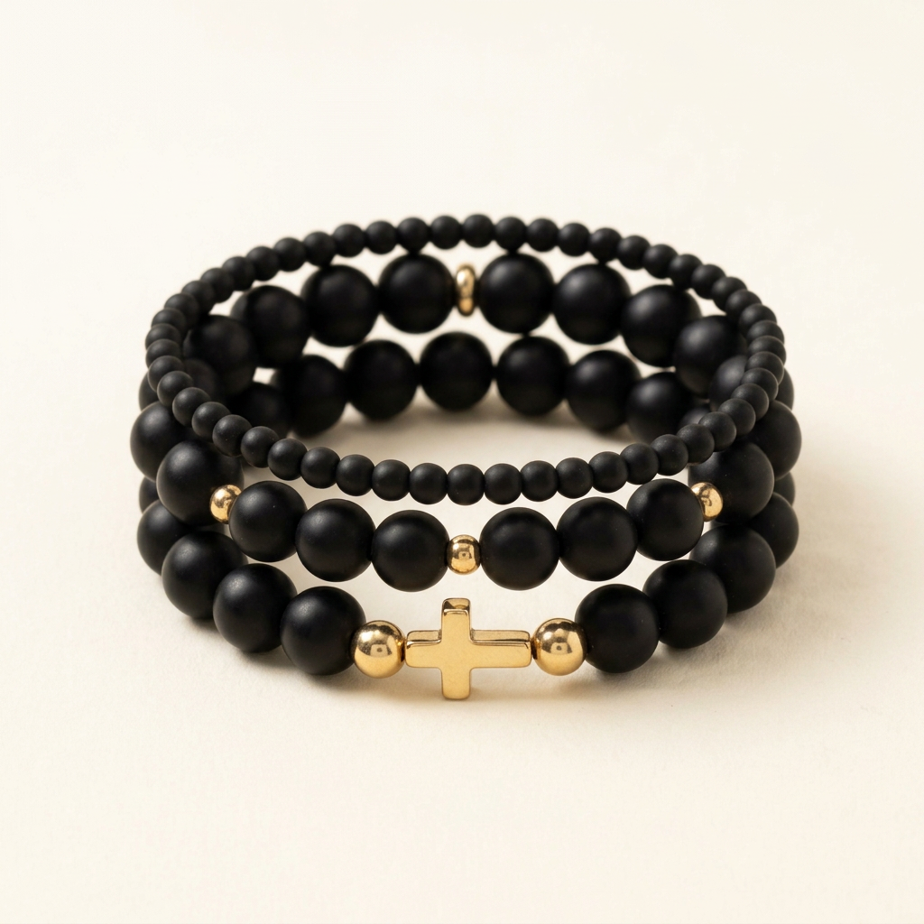 Three handmade black Onyx bracelets arranged in a three-tier stack with gold colored accents and a gold colored cross on an ivory background