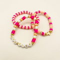 Three pink, peach, and cream clay bead bracelets with gold-tone accents, a cross, and WHO ELSE lettering on an ivory background