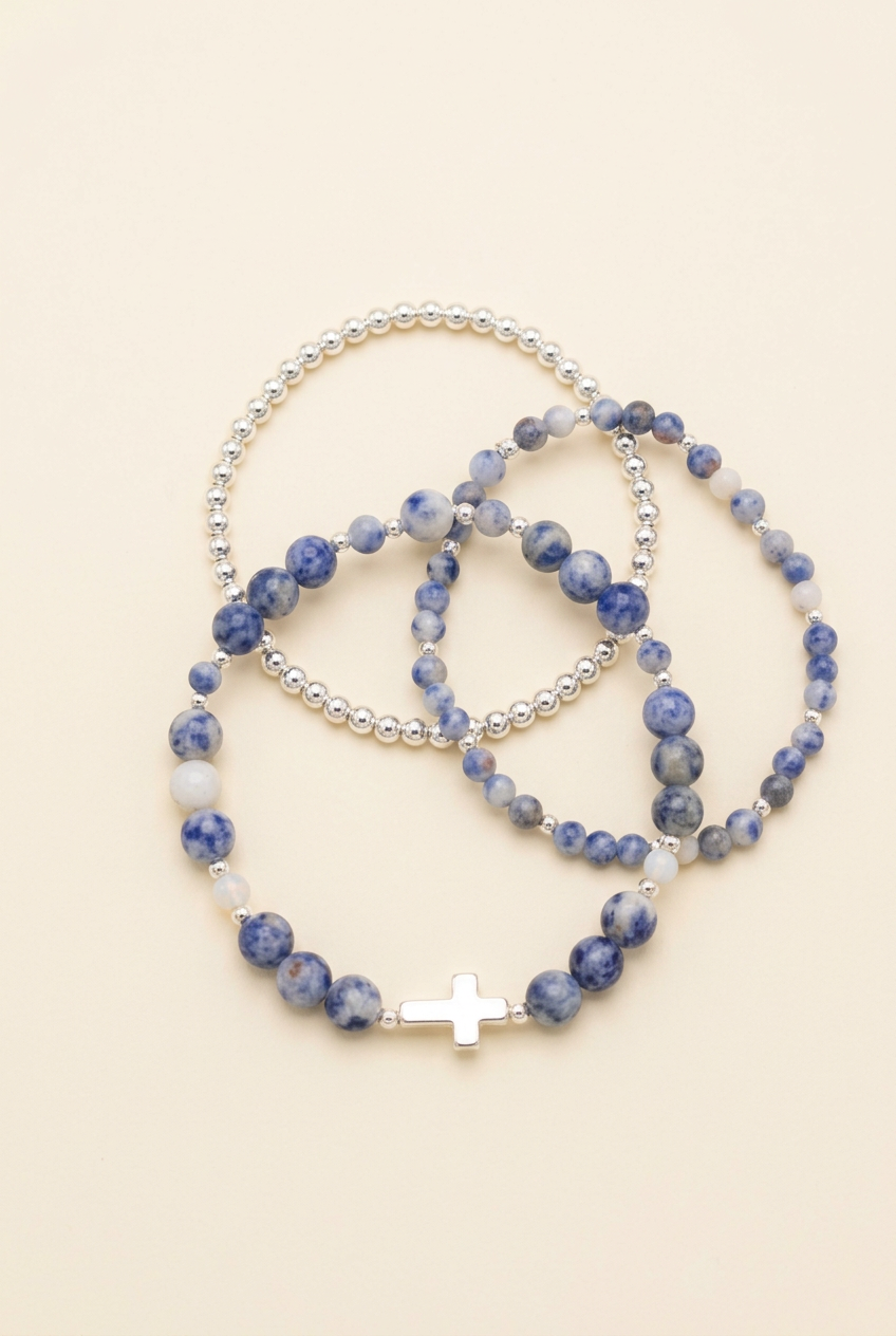 Three handmade blue jasper and silver-accented bracelets with a cross on a warm ivory background