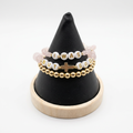 Three stacked pale pink and gold-tone beaded bracelets with white letter beads and a cross detail on a black display stand.