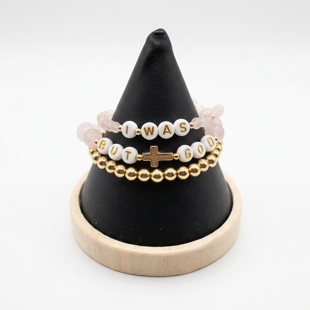 Three stacked pale pink and gold-tone beaded bracelets with white letter beads and a cross detail on a black display stand.
